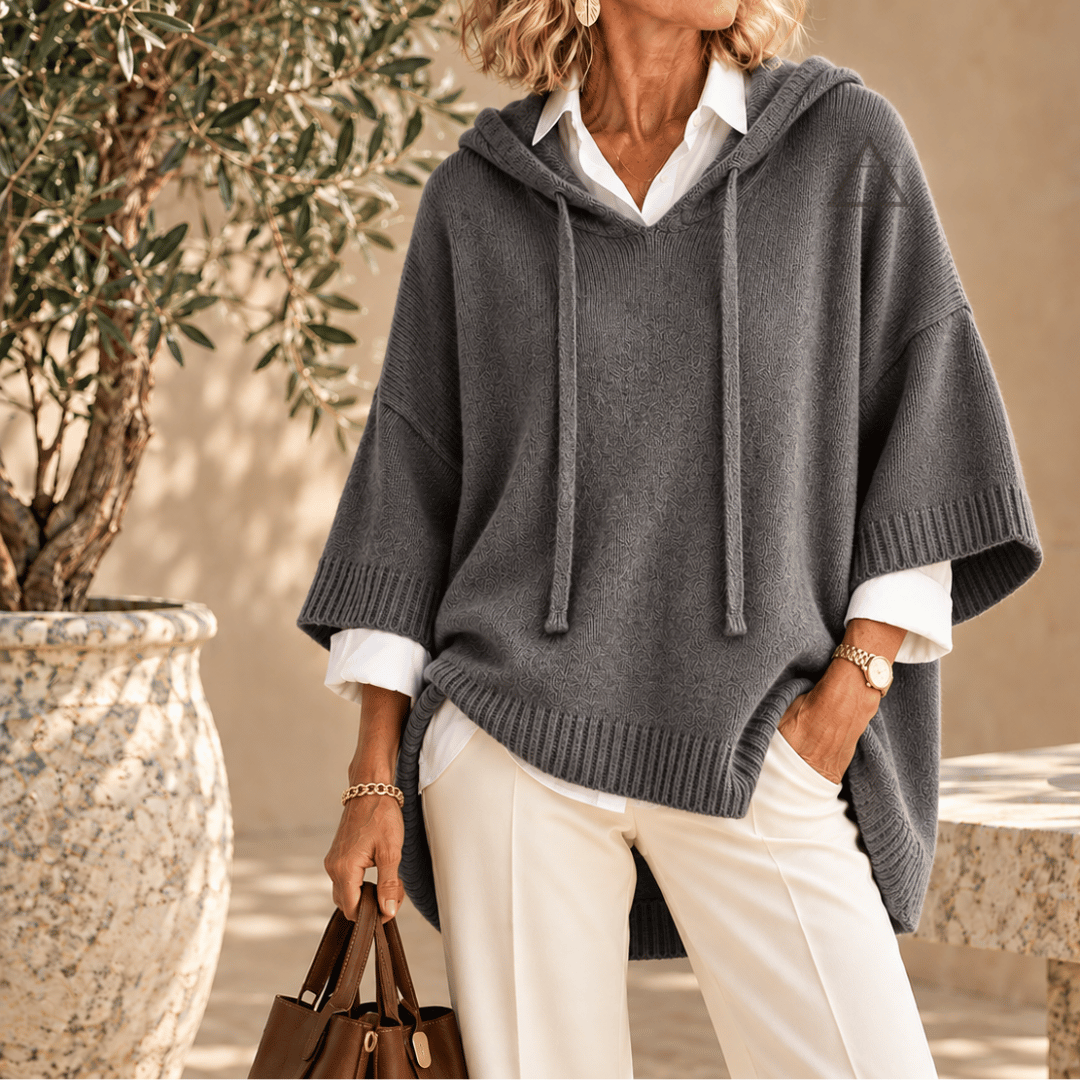 Poncho Clémence en maille gris anthracite, à capuche et finitions côtelées, porté sur une chemise blanche avec un pantalon blanc.
