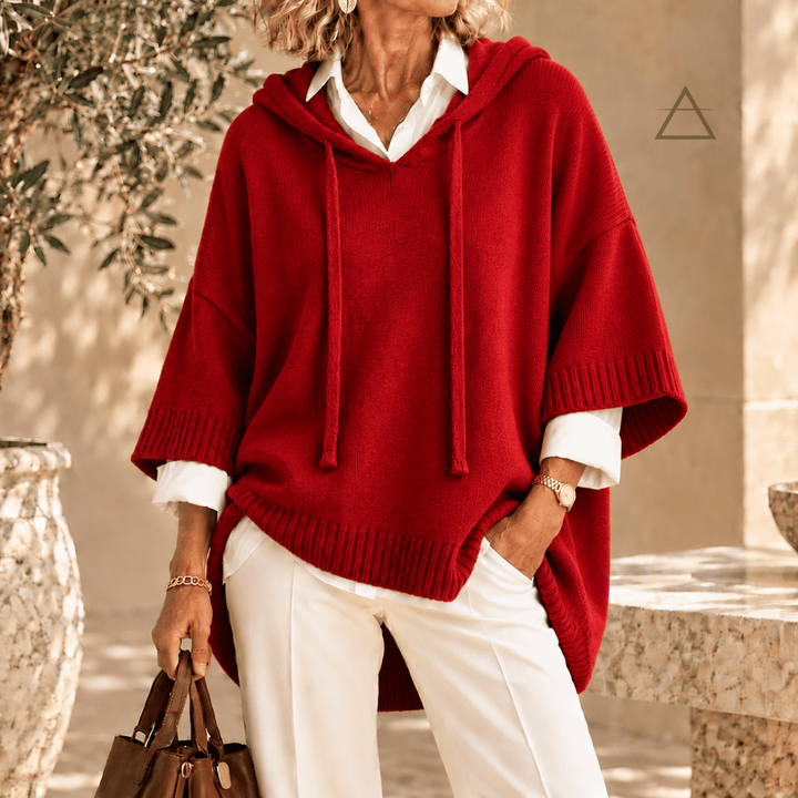 Poncho Clémence en maille rouge cardinal à capuche et finitions côtelées, porté sur une chemise blanche avec un pantalon blanc.
