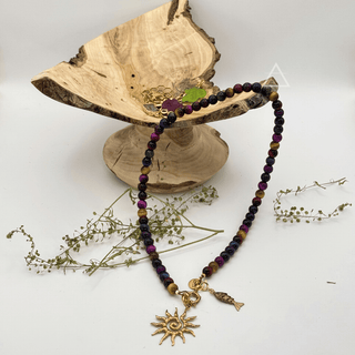 Collier SOLSTICE à perles multicolores et pendentifs soleil et poisson dorés, disposé près d’un présentoir en bois.