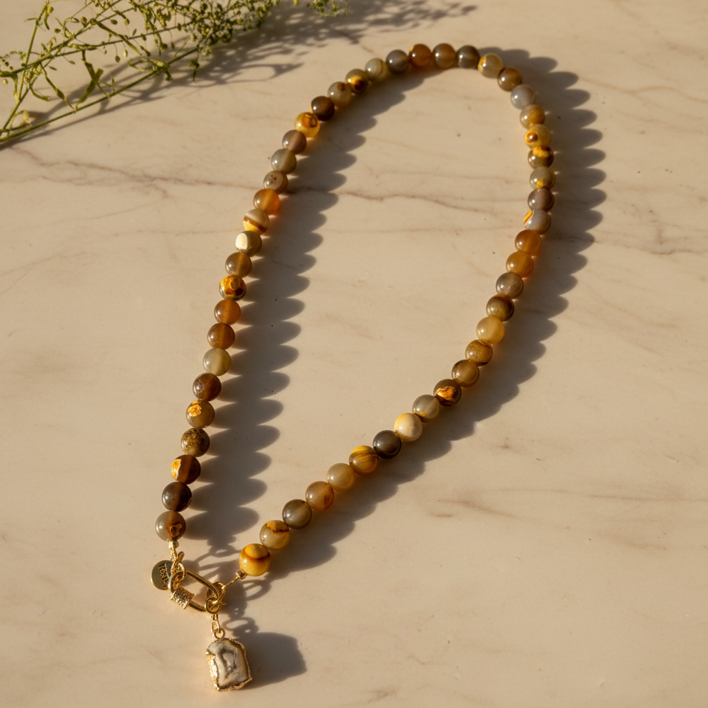 Collier Unspoken aux perles brunes, miel et grises, avec pendentif en agate, posé sur un fond clair en lumière chaude.